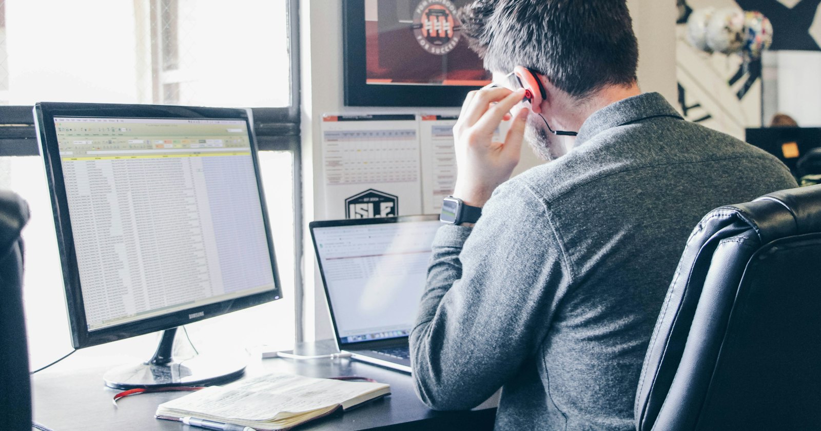 A person on the phone at an office desk, with a spreadsheet on the monitor